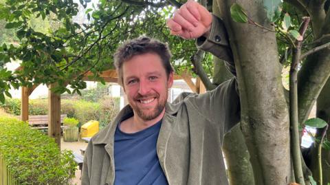 Charlie standing in front of a tree in a garden. He is wearing a beige jacket.