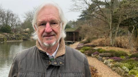 Councillor Roly Cronshaw has white hair, a beard and is wearing glasses. He is also wearing a dark brown jacket and is smiling at the camera. He is standing in front of a pond, close to trees and flowers.