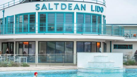 A white and turquoise Art Deco lido, with Saltdean Lido written in lettering across its frontage. A woman in a red bathing cap swims in the pool in front of it. 