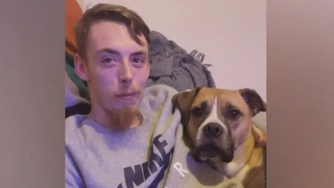 Charlie Greaves smiles at the camera while hugging a dog. He is wearing a grey sweatshirt.
