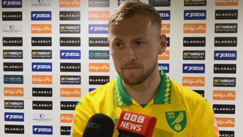 Norwich City player Harry Darling wearing club shirt standing in front of sponsors logo board
