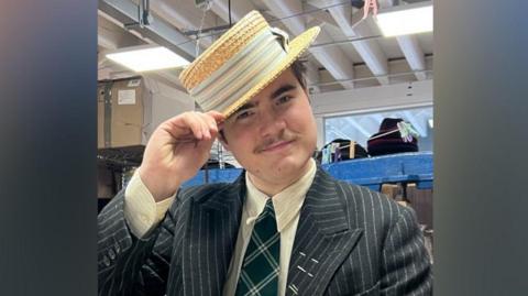 A person in a dark pinstripe suit, cream shirt, and green plaid tie stands indoors holding a straw boater hat. Behind them are shelves filled with hats and boxes in a workshop‑like space with exposed beams and fluorescent lighting.