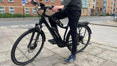 A generic photo of a person on a pavement astride a black e-bike, showing their legs, one of which is on a pedal. They are wearing dark trousers and blue trainers and there are red brick flats and a road in the background