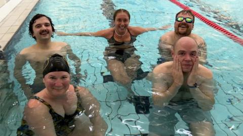 The five challengers are smiling while submerged up to their shoulders in the swimming pool. Dan's hands are around his face and he looks worried.