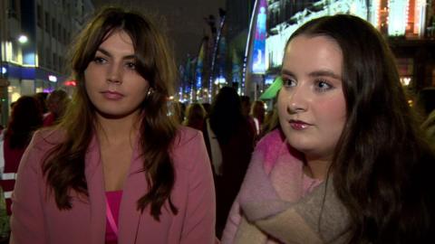 two women stand stand outside City Hall. The women on the left has brown hair, wears a pink blazer with a pink top underneath. The other has on a Pink and grey scarf with a pink hi-viz jacket, she has brown hair