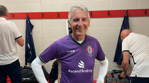 Drew Warren in his football kit smiling for the camera at a dressing room. Two other men can be seen behind him.