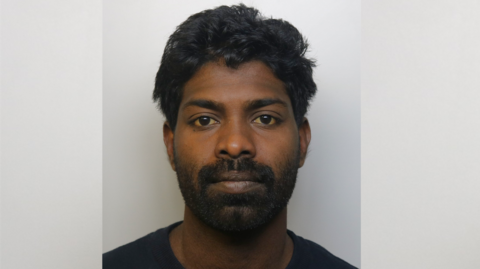 A police mugshot of Manoj Chinthathira wearing a black t-shirt and standing against a grey background. He has brown eyes, a full black beard and thick wavy black hair.