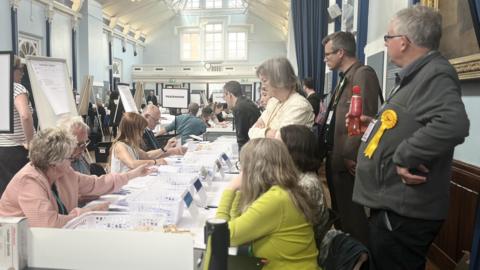 Delegates stand up and observe as ballot papers are counted and sorted by people sat at a long table. They are in a large hall with high ceilings.