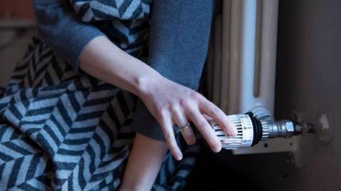 A close-up of someone with their back to a radiator and a blanket over them, turning to adjust the temperature on the dial.