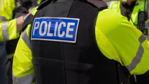 This picture shows three police officers in uniform. None of the officers are identifiable. They are wearing a high-vis jacket with a black short-sleeved jacket. On the back of one of their jackets it says Police