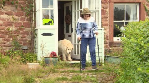 Carol Smithers walks out of a door of her home, holding two walking sticks. She is followed by a golden retriever, which is in the doorway behind her.