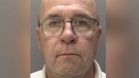 A man with a bald head and stubble beard wearing glasses and a cream T-shirt. He is staring into the camera for a police mugshot.