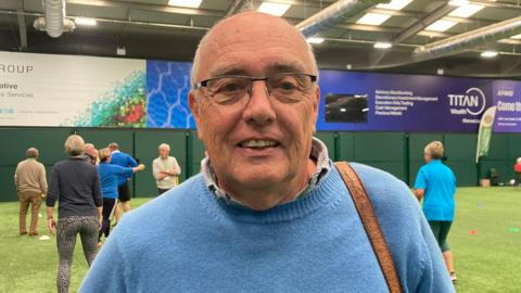 A mid-shot of John Adams. He has grey hair, wears glasses, and has a blue jumper. In the background are the carers and others playing sport.