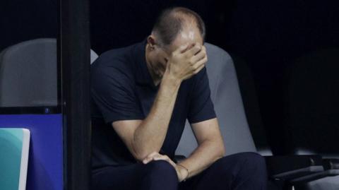Thomas Tuchel looks down and scratches his head while sitting on the bench after England lost to Argentina