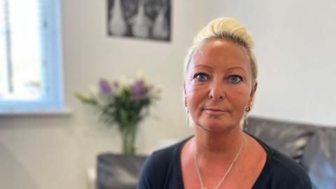 Charlotte Charles sits on a black sofa. She is wearing a black T-shirt and her blonde hair is up. 