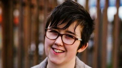 Lyra Mckee is smiling and wearing glasses in a grey coat