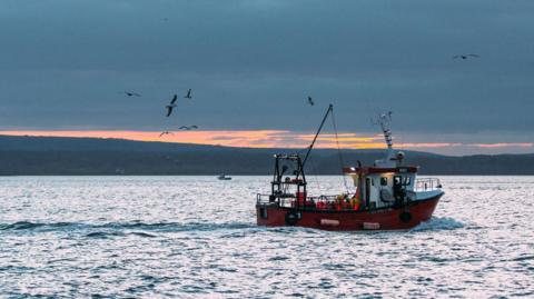 Sandbanks, Dorset - November 24, 2020: A small red fishing trawler with fisherman on deck returning to Poole harbour with their catch surrounded by seagulls with light waves at dusk.