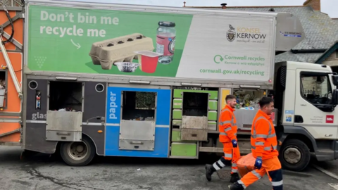 Two waste disposal staff wearing high viz jackets, walking in front of a Cornwall Council recycling lorry.