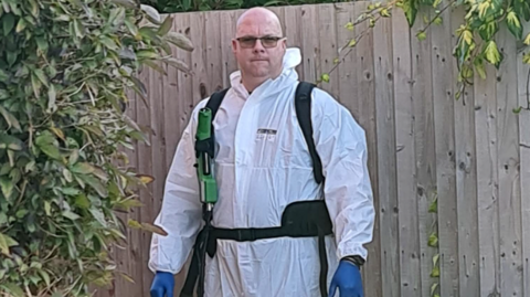 A man in a white suit and blue gloves, with a spray gun on a strap over one of his shoulders
