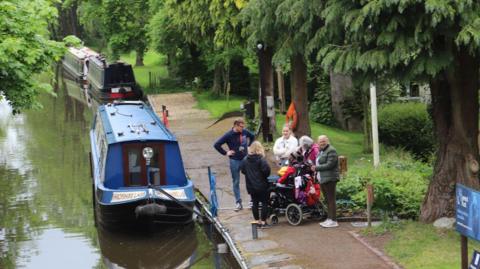 A young person in a wheelchair, surrounded by five adults on a canal towpath with a blue canal boat in the foreground and two other canal boats in the distance