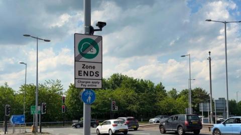 A busy urban road junction on a partly cloudy day. In the foreground, there is a tall grey pole positioned slightly to the left of the center. Attached to this pole is a rectangular road sign with a green circular symbol that contains a cloud-like shape and the letter “C” A diagonal black line crosses through the symbol, indicating the end of a designated zone. Beneath the symbol, the sign clearly reads “Zone ENDS” in bold black letters, followed by smaller text stating “Charges apply Pay online”. Below this sign, there is a smaller circular blue sign showing a white bicycle and a pedestrian, indicating a shared path for cyclists and pedestrians.