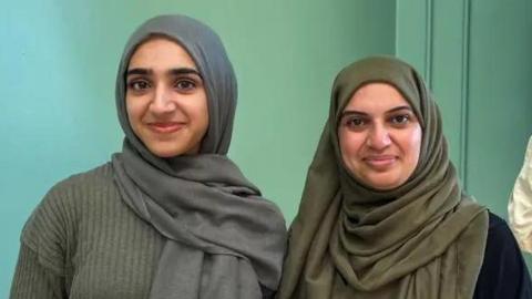 Husna, wearing a grey jumper and a grey headscarf, with her mum, in a green  headscarf, smiling for the camera.