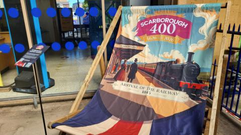 A giant decorative deck chair stands outside a building with glass doors, its fabric printed with colourful artwork celebrating Scarborough’s 400th anniversary. The design shows a steam locomotive arriving at a station with people in period clothing, alongside text marking historical dates.