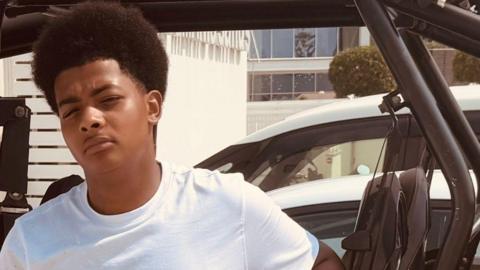 A teenage boy leaning against a car, wearing a white top