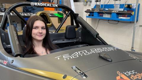 A young woman sat in the cockpit of a sports car in a mechanics shop