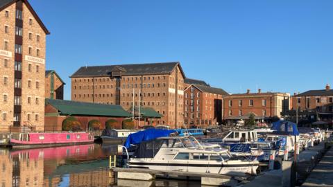 A photo of Gloucester Docks. There are a number of large orange brick warehouse buildings. In the docks there are a number of small blue and white speedboats moored up, and several canal boats behind them.