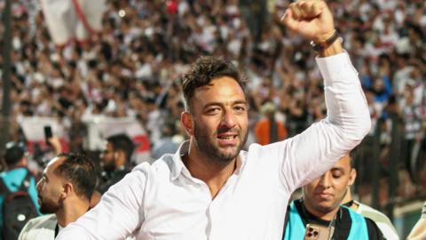 Mido, wearing a white shirt with the neck open, raises his left fist in the air at a football match as photographers in turquoise bids are seen behind him. In the far background a blurry crowd pushes up against a fence