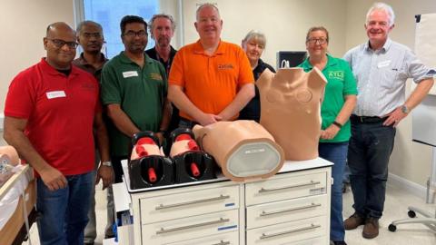 A group of medical instructors in different coloured polo shirts in a hospital room, with training mannequins that resemble torsos placed on a table in the centre of the room.