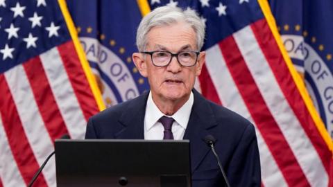 Jerome Powell, a man with grey hair and black square glasses, speaks at a podium in front of American flags and Federal Reserve flags, wearing a dark suit and purple tie