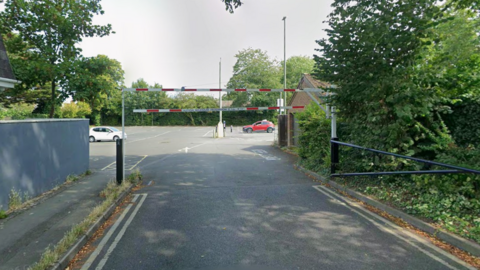 A road leading to a large car park, which is behind a red and white height restriction barrier.
