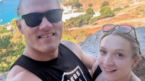Jess - a short blonde woman in her 20s - stands to the right of her partner Jake who is wearing a black tank top and has his arm around Jess. They both are looking upwards to the camera and smiling. They're standing on a hill with a Mediterranean-looking coastal background behind them in this holiday snap. 