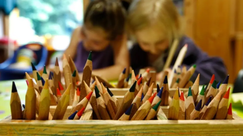 Boxes of coloured pencils sit in the foreground of the image with two children writing and slightly blurred in the background