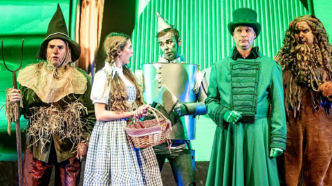 Five actors are standing on stage dressed in costume. They are characters from the Wizard of Oz.