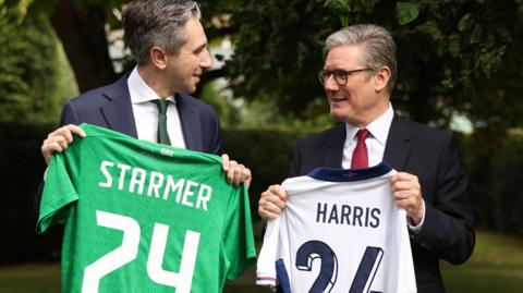 Simon Harris holding a green football jersey with the word 'Starmer' on the back, standing beside Sir Keir Starmer holding a white football top with the word 'Harris' on the back