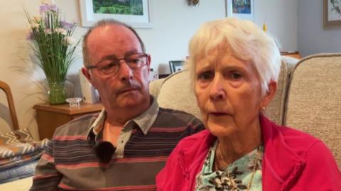 George and Jill Bristow sit on a sofa in a living room. George is wearing a striped shirt and has glasses. Jill has white hair and is wearing a red jacket over a floral top.