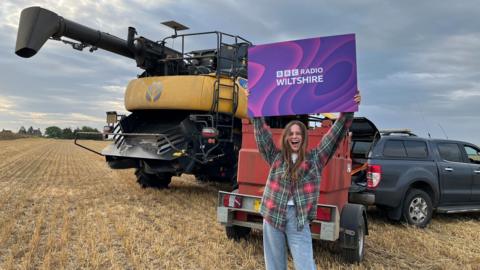 Farmwatch Gabrielle Aplin singer and sheep farmer holds a purple sign saying 'BBC Radio Wiltshire' in front of farming equipment