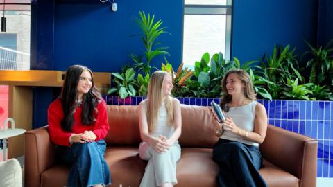 Three students sat on a brown sofa. The student on the right is showing the other two a cocktail shaker