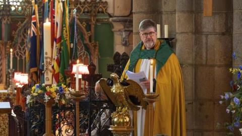 Tim Barker standing at the lectern of a church holding a sheet of paper while leading a service. He is wearing yellow and green vestments. There are candles ad flowers in the church
