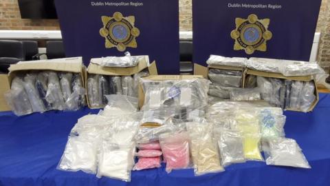 Bags of suspected drugs lined up on a table with posters of An Garda Síochána's logo hung up behind it