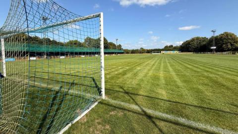 A stock image of a football pitch with green grass and a goal with blue and white netting. There are stands and trees in the far distance.