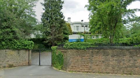 A wall with a gate in the middle with a white building among trees behind it, and a sign saying Nottinghamshire Healthcare NHS Trust Mandala Centre on the wall