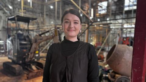 Woman with dark hair tied back, smiling and wearing a black top in front of a construction site 