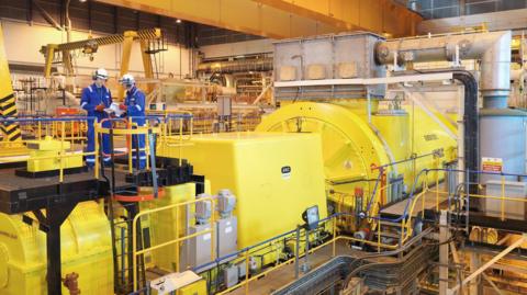 Two workers wearing helmets and blue overalls are talking to each other on a metal platform above a yellow generator. The long, yellow metal tube takes up most of the space. There are various pipes and monitors coming off it. "Generator 2" is written on the side.