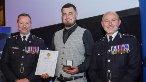 A man with dark brown hair and short beard, wearing a grey waistcoat and black shirt and tie, holding a police bravery award certificate and a medal in the other hand. He is flanked on either side by two senior police officers.