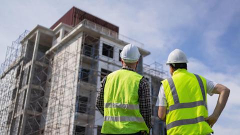 Two builders in high visibility jackets and white hard hats look towards a tall set of unfinished flats with scaffolding around it.