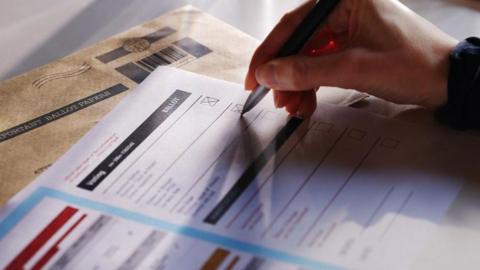 A close-up shot of a ballot paper with an unknown person's right hand holding a pen and marking an X on the paper. Only the hand is visible.